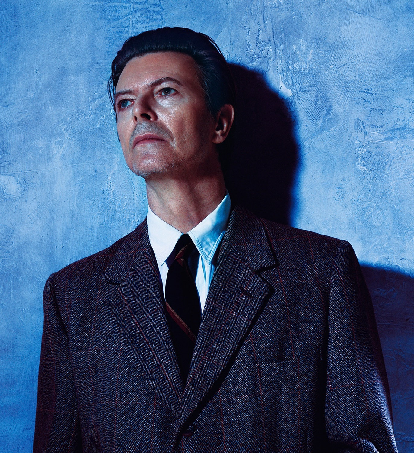 David Bowie in a dark tweed suit and tie, gazing upward against a mottled blue wall.