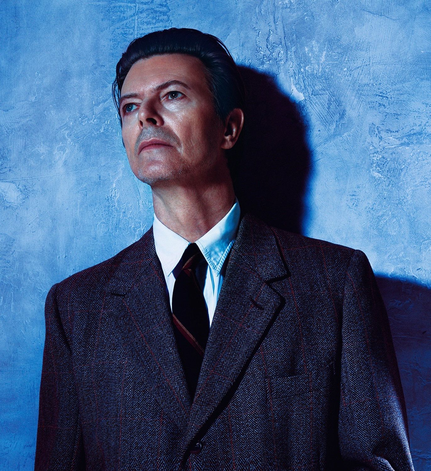 David Bowie in a dark tweed suit and tie, gazing upward against a mottled blue wall.